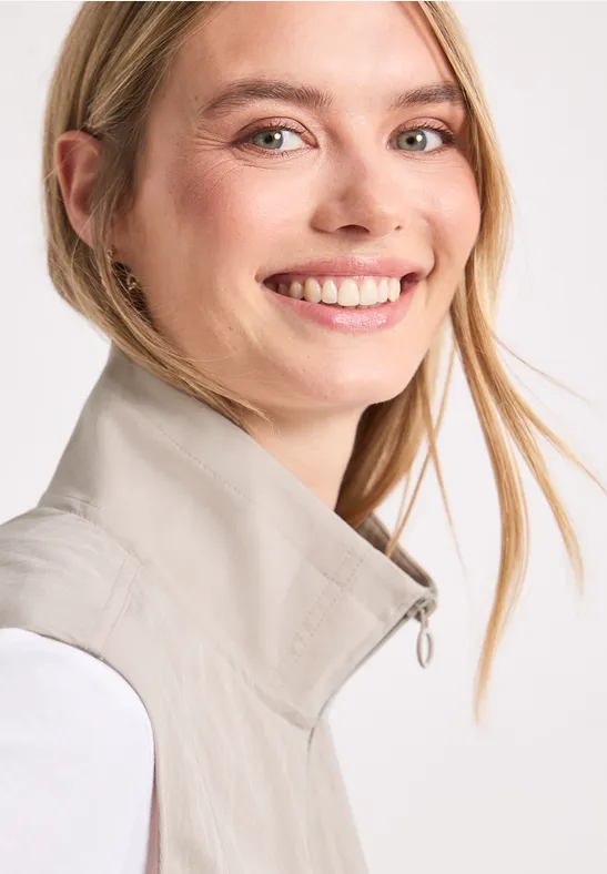 Eine lächelnde Frau mit blondem Haar trägt eine helle Weste vor weißem Hintergrund.