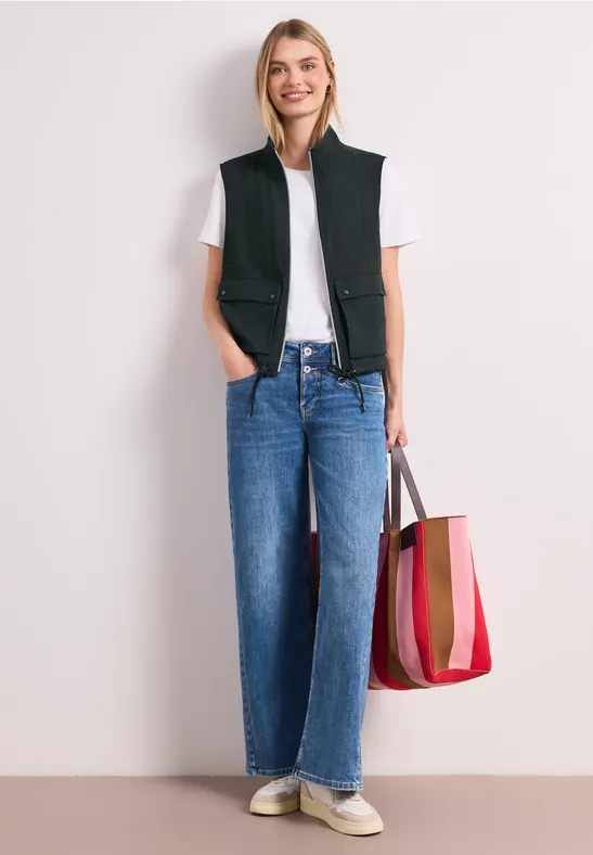Femme en T-shirt blanc, gilet noir, jean bleu droit, baskets blanches et sac rouge à rayures