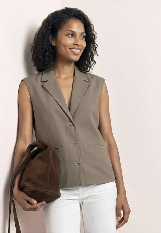 Femme portant un blazer sans manches taupe, pantalon blanc et sac marron.