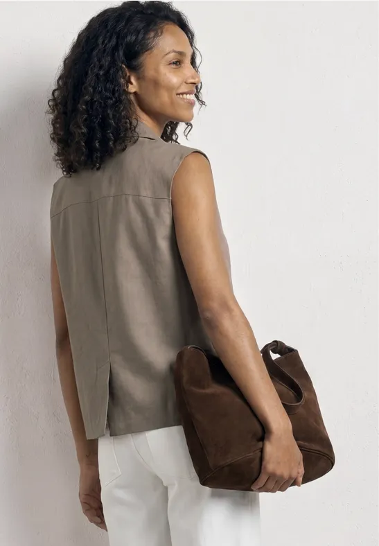 Femme vue de dos en top sans manches couleur terre, pantalon blanc, tenant un sac en daim marron.
