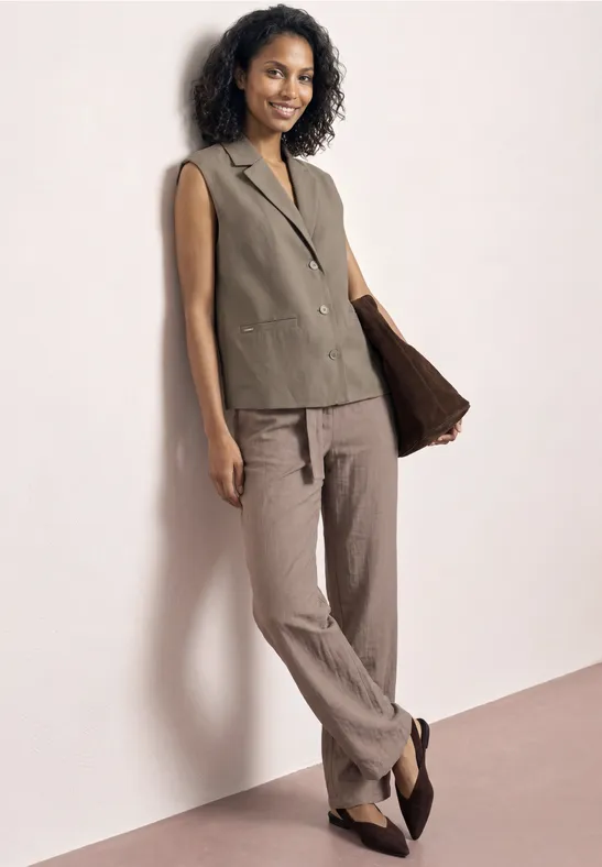Femme appuyée contre un mur blanc, veste olive sans manches, pantalon beige et ballerines noires.