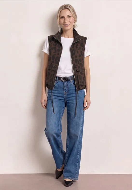 Femme en t-shirt blanc, gilet sans manches marron à motif, jean évasé bleu et chaussures noires.