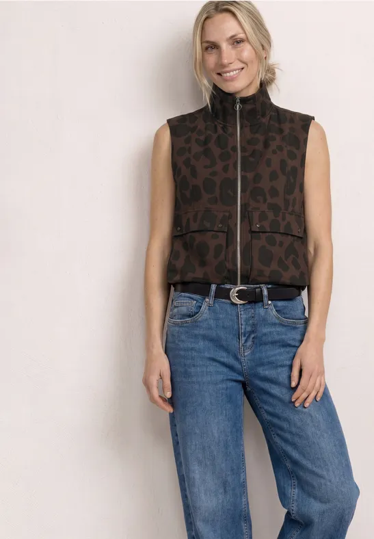 Femme en gilet sans manches léopard à fermeture éclair et jeans bleus sur fond clair.