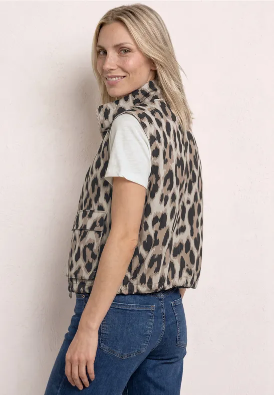 Femme de profil en jean, t-shirt blanc et gilet léopard devant un mur clair