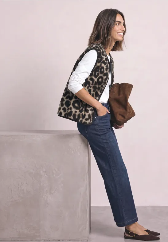 Eine lächelnde Frau mit Leopardenmuster-Weste, weißem Shirt und Jeans lehnt an einem Block und hält eine braune Jacke.