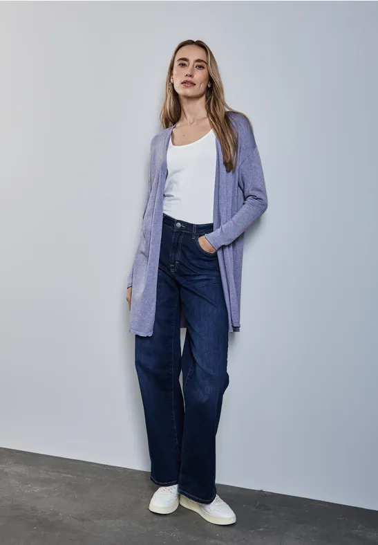 Frau in lila Cardigan, weißem Shirt und Jeans steht an einer Wand.