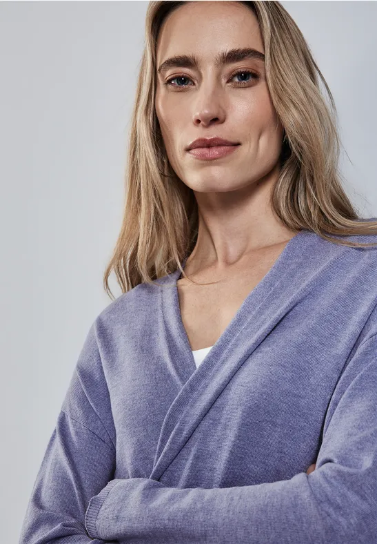 Frau mit langen blonden Haaren und lila Pullover schaut selbstbewusst.