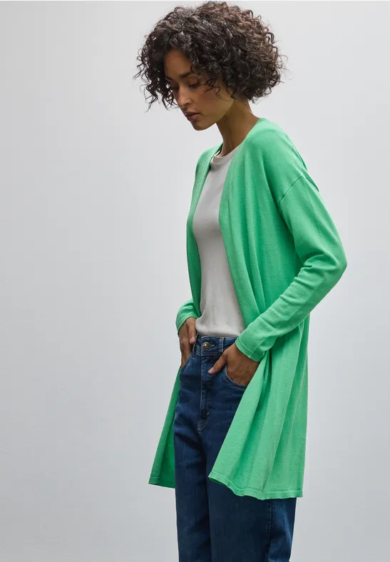 Frau in grünem Cardigan, weißem Shirt und Jeans, lehnt lässig an der Wand.