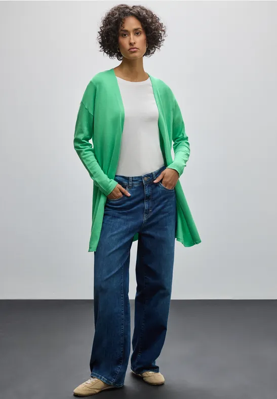 Frau mit grünem Cardigan, weißem Shirt, Jeans und Sneakers läuft seitlich.