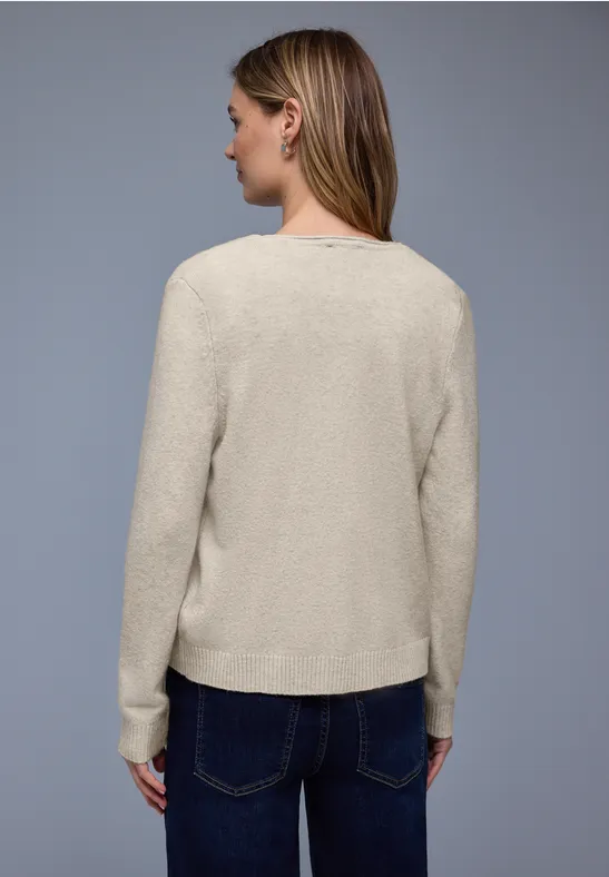 Rückansicht einer Person mit beigem Strickpullover, weißer Bluse und dunkelblauen Jeans vor grauem Hintergrund.