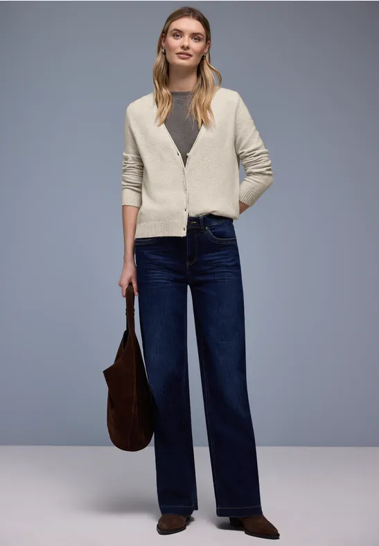 Eine Frau in cremefarbenem Cardigan, grauem Shirt und weiten, dunkelblauen Jeans hält eine braune Tasche.