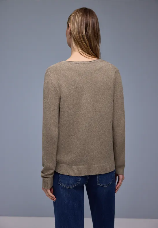 Eine Frau von hinten in einem taupefarbenen Pullover und blauen Jeans vor grauem Hintergrund.