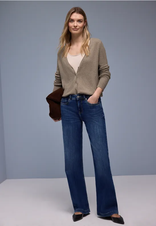 Frau in grünem Cardigan, weißem Top, weiten Jeans, spitzen Schuhen und Clutch vor grauem Hintergrund.
