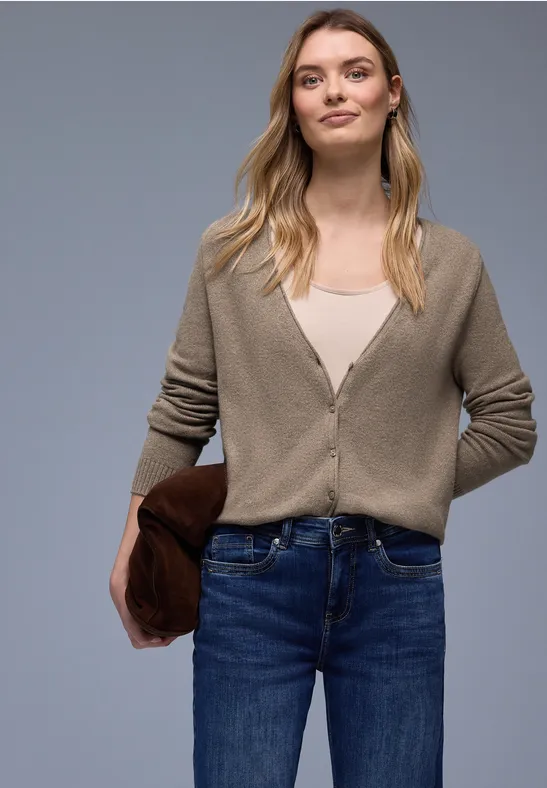 Eine Frau trägt beige Strickjacke, weißes Top, blaue Jeans und hält eine braune Clutch vor grauem Hintergrund.