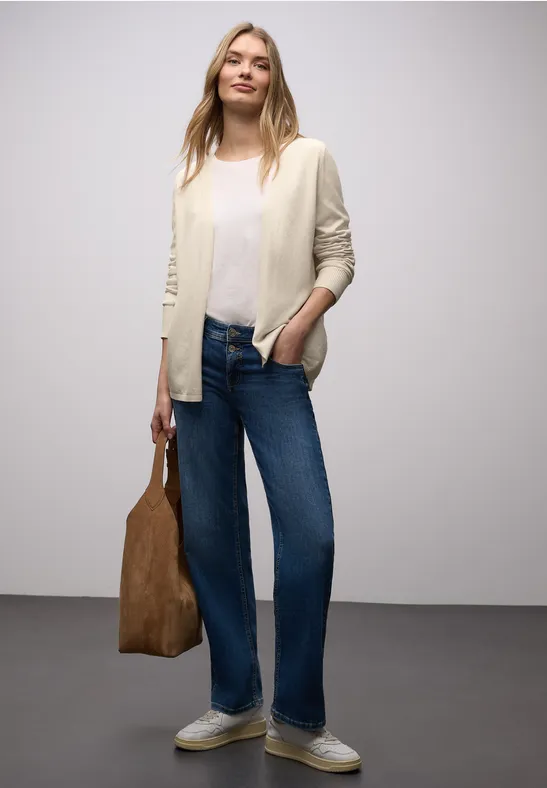 Vrouw met witte blouse, beige vest, wijde jeans, witte sneakers en bruine tas voor een grijze achtergrond.