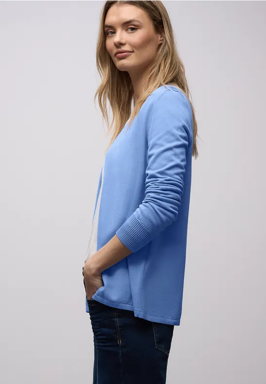 Frau im Profil mit hellblauem Cardigan, weißem Shirt und dunklen Jeans vor neutralem Hintergrund.
