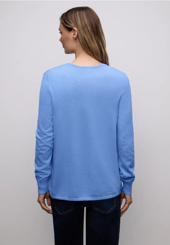 Eine Person steht mit dem Rücken zur Kamera und trägt einen hellblauen Pullover und eine dunkle Hose.