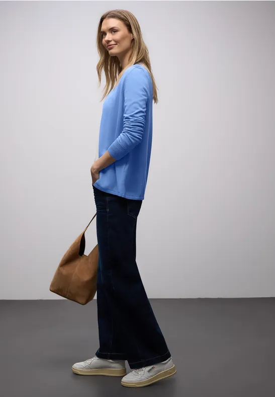 Person im Profil trägt hellblauen Pullover, weite dunkle Jeans, weiße Sneaker und braune Tasche.