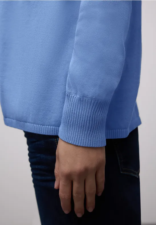 Eine Person trägt ein hellblaues Sweatshirt und dunkle Jeans. Arm und Hand sind sichtbar.