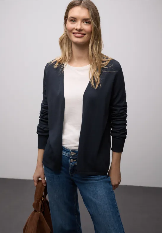 Eine Frau trägt eine dunkelblaue Strickjacke, weißes T-Shirt, Jeans und hält eine braune Tasche.