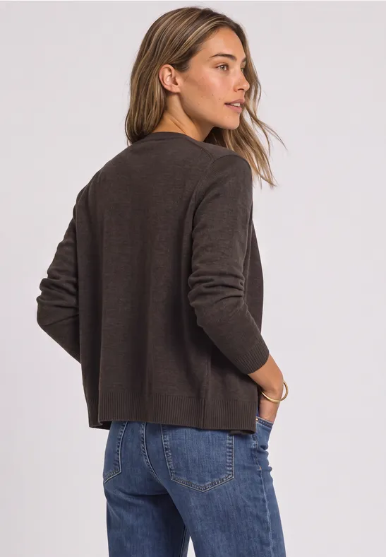 Frau steht seitlich, trägt braunen Pullover und Jeans, legt Hände in die Taschen und blickt über die Schulter.
