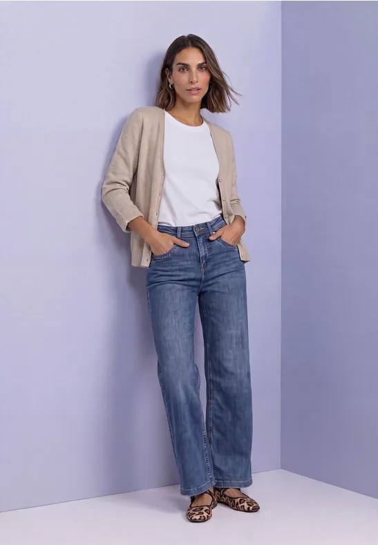 Eine Frau mit weißem T-Shirt, beigem Cardigan, weiten Jeans und Leo-Sandalen vor violettem Hintergrund.