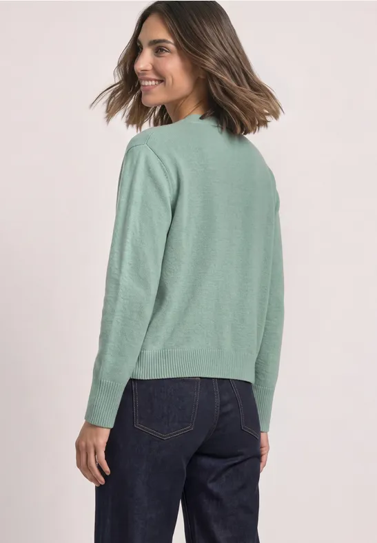 Une femme porte un pull vert clair et un jean foncé, souriante, regard par-dessus son épaule.