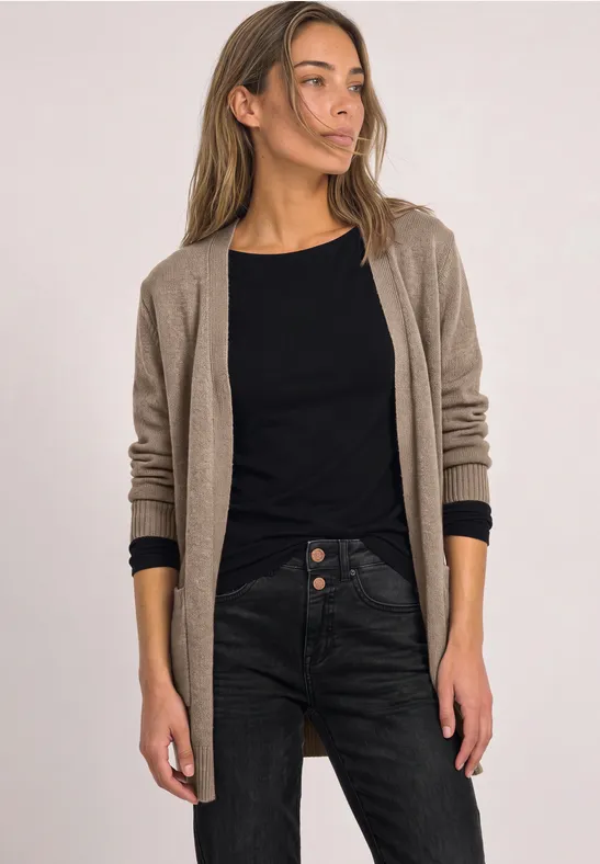 Een vrouw draagt een lange beige cardigan, een zwart shirt en donkere jeans, staand voor een lichte achtergrond.