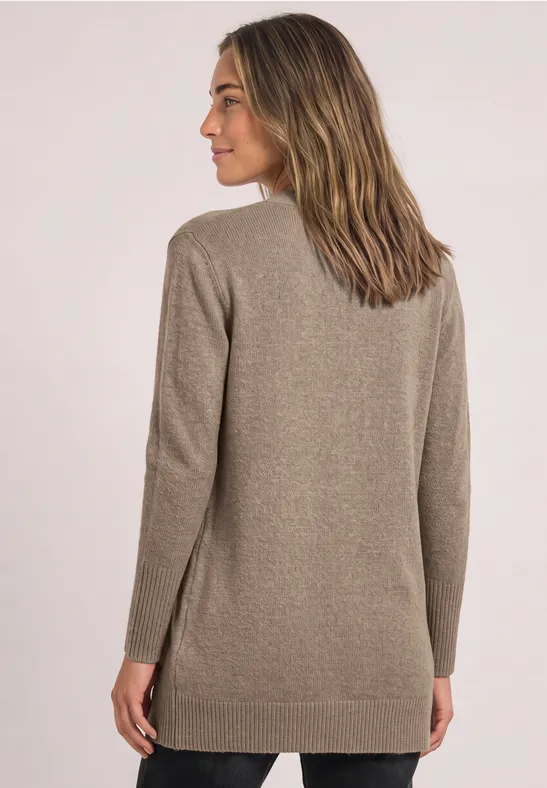 Vrouw in een lang taupekleurig gebreid vest, half zijwaarts staand voor een lichte achtergrond, kijkt naar links.