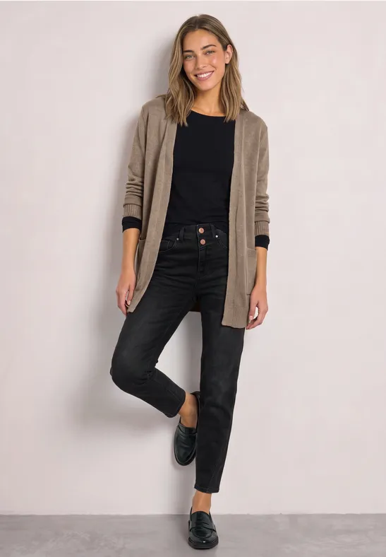 Jonge vrouw in een beige vest, zwart shirt, donkere spijkerbroek en zwarte loafers voor lichte achtergrond.