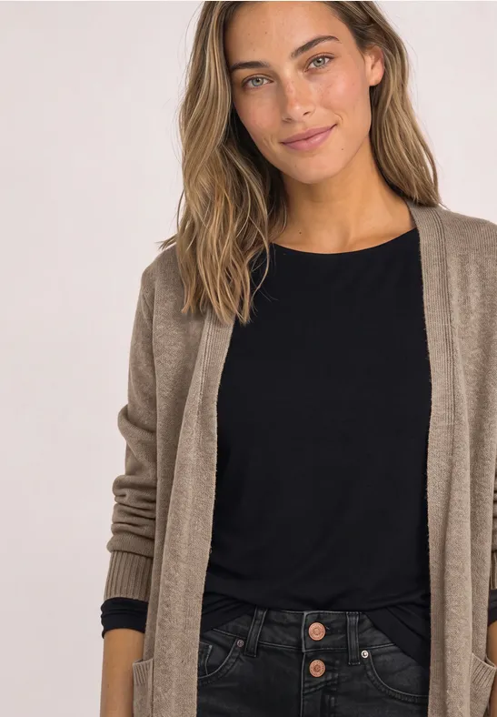Een glimlachende vrouw met bruin haar draagt een zwart shirt en een beige vest tegen een lichte achtergrond.