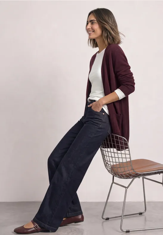 Frau in burgunderrotem Cardigan, weißem Shirt und weiten Jeans lehnt sich lächelnd an Stuhl.