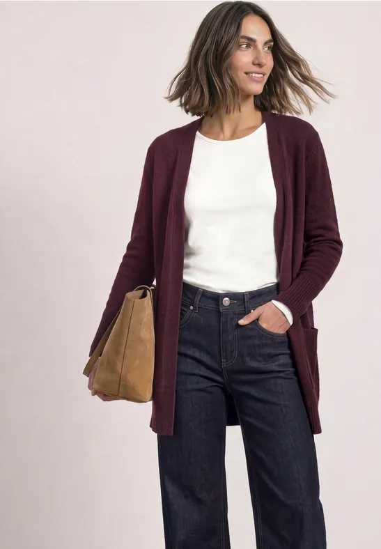 Frau im weinroten Cardigan über weißem Shirt, dunklen Jeans und beiger Tasche, lächelnd, natürlicher Look.