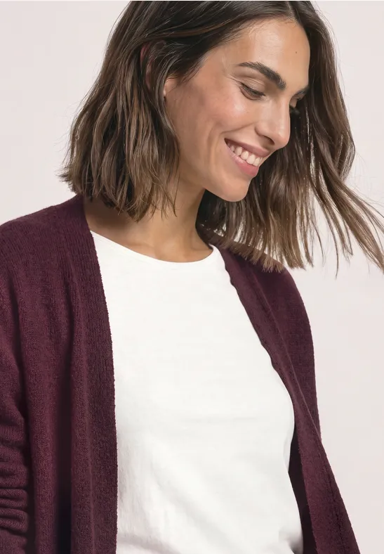 Eine lächelnde Frau mit weißem T-Shirt und bordeauxrotem Cardigan.