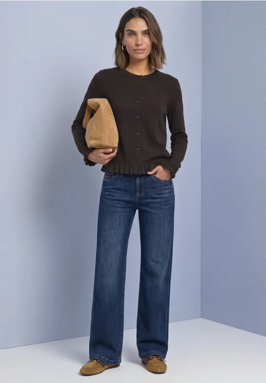 Vrouw in een bruin trui en wijde jeans houdt een bruine tas vast voor een lichtblauwe achtergrond.