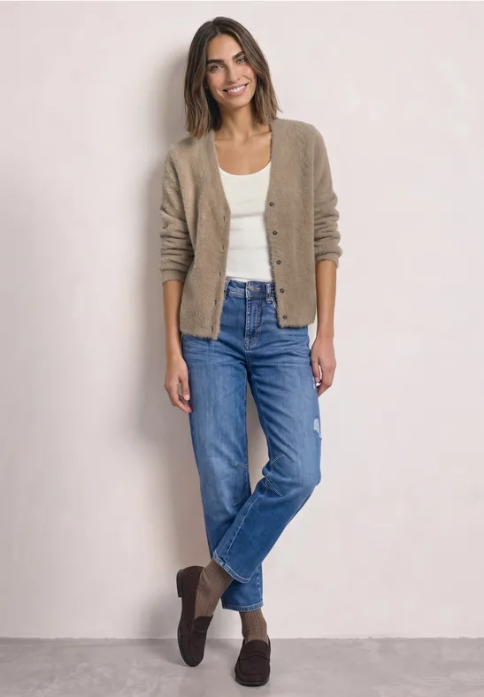 Vrouw in spijkerbroek, wit shirt en beige vest, ontspannen poserend voor een lichte achtergrond.