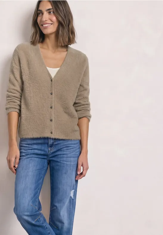 Een vrouw draagt een beige vest en blauwe spijkerbroek, staand glimlachend voor een lichte achtergrond.