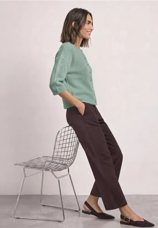 Femme appuyée sur chaise métal, pull vert menthe, pantalon marron ample et mocassins bicolores.