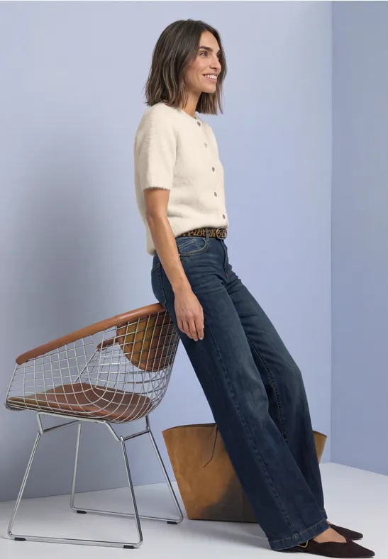 Vrouw in beige T-shirt en wijde jeans bij een draadstoel en houten blok tegen een lichte achtergrond.