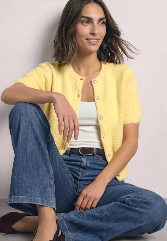 Een vrouw zit in kleermakerszit, draagt een geel vest, een wit shirt, blauwe jeans en lacht.