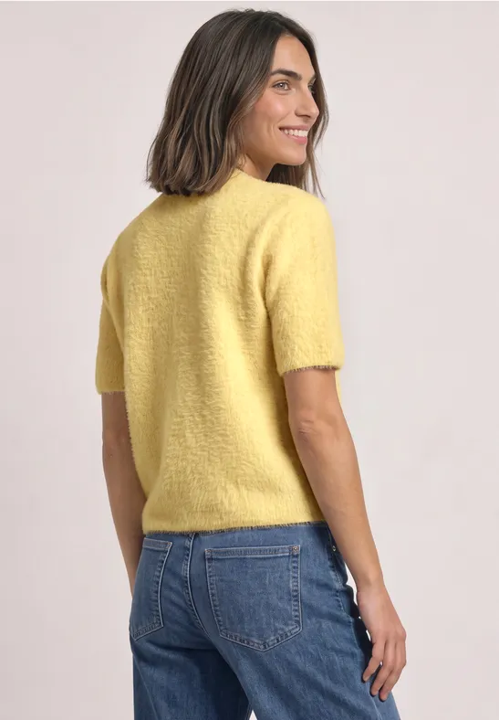 Vrouw in geel kort mouw trui en jeans, van achteren, zijwaarts kijkend, glimlachend.