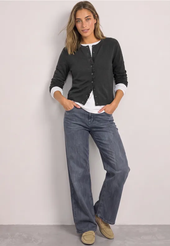 Femme en gilet noir, t-shirt blanc et jean large, appuyée de façon décontractée contre un mur gris.