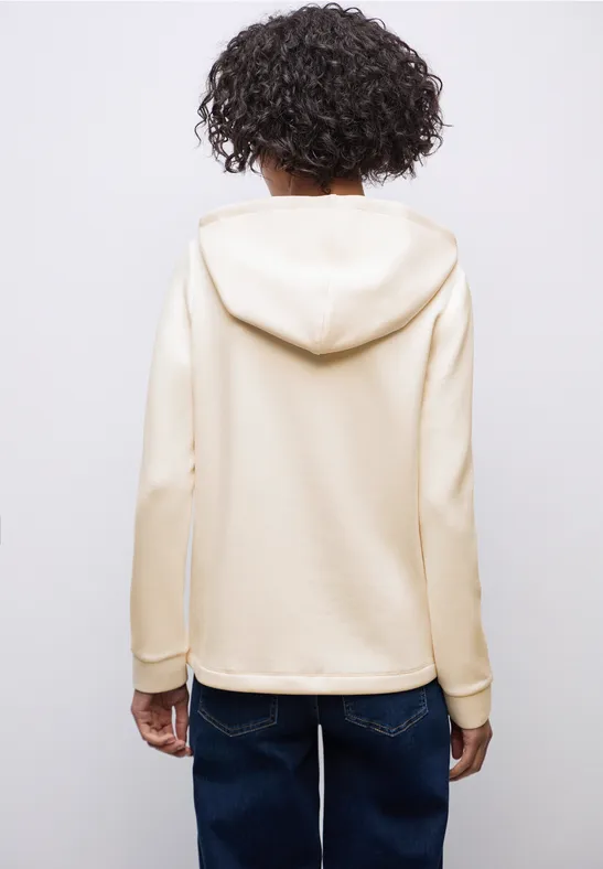 Frau von hinten mit cremefarbenem Hoodie und Jeans vor grauem Hintergrund.