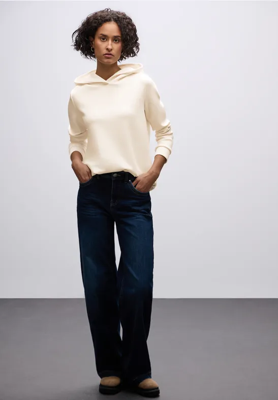 Frau in beiger Kapuzenjacke und weiter Jeans lehnt an einer grauen Wand.