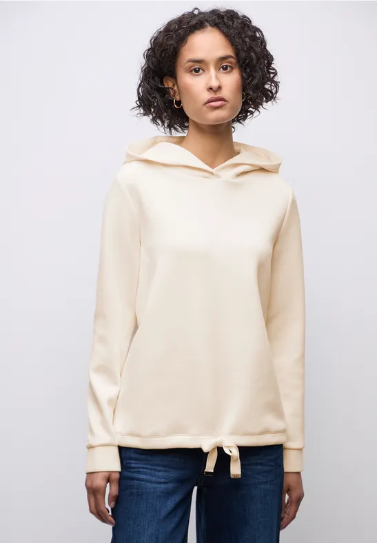 Frau mit lockigem Haar trägt cremefarbenen Kapuzenpullover und Jeans.