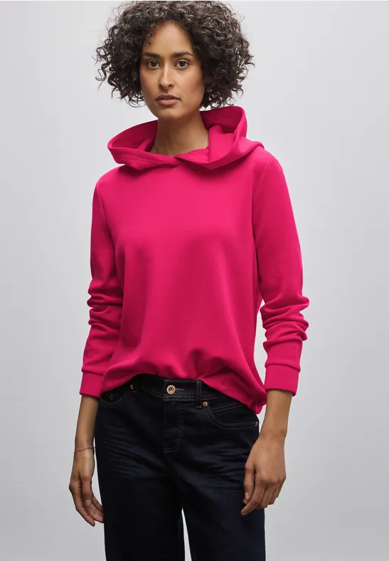 Frau mit blondem Haar trägt einen pinken Hoodie und Jeans.