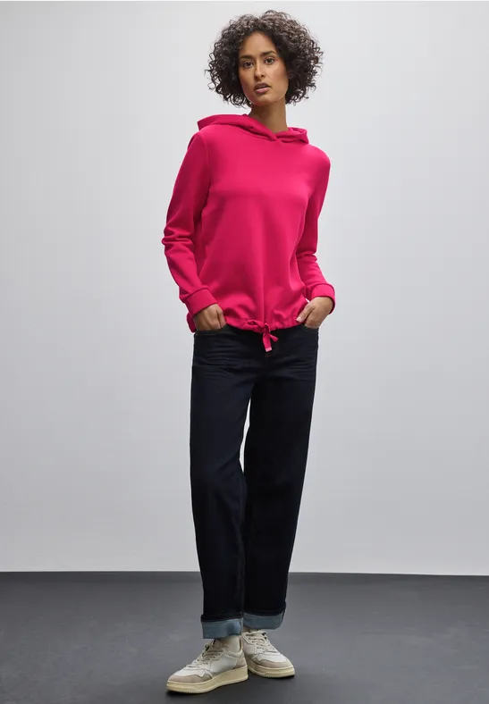 Frau in pinkem Hoodie, blauer Jeans und weißen Sneakers vor grauer Wand.