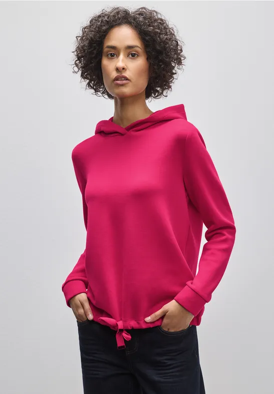 Frau mit lockigem Haar trägt pinken Kapuzenpullover und dunkle Hose.