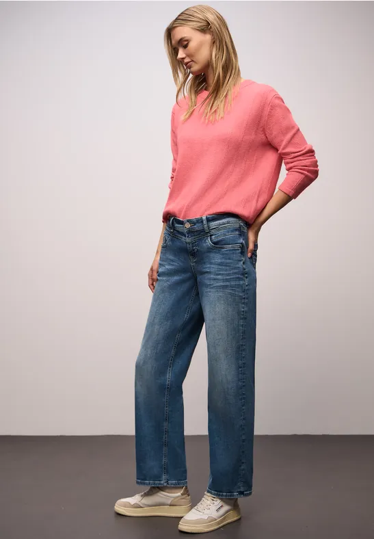 Frau in pinkem Pullover, blauen, weiten Jeans und weißen Sneakern steht locker mit einer Hand in der Hosentasche.