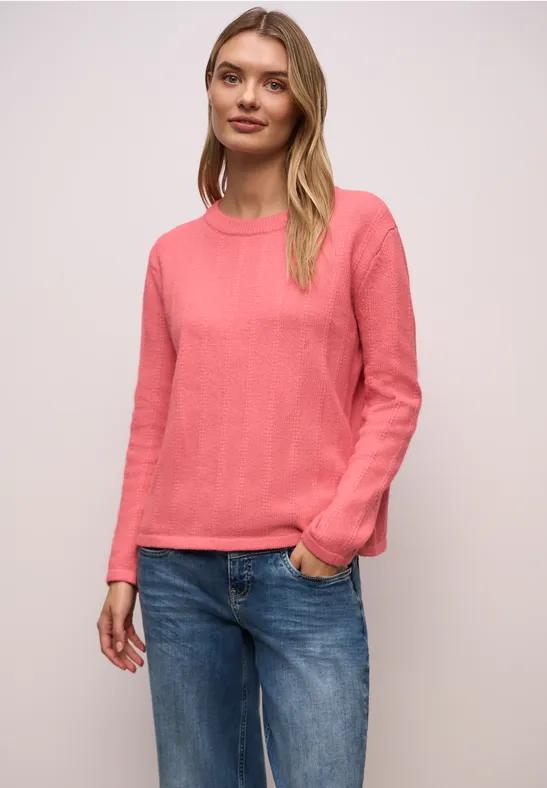 Frau in pinkem Pullover und blauen Jeans, steht lässig mit einer Hand in der Tasche vor hellem Hintergrund.